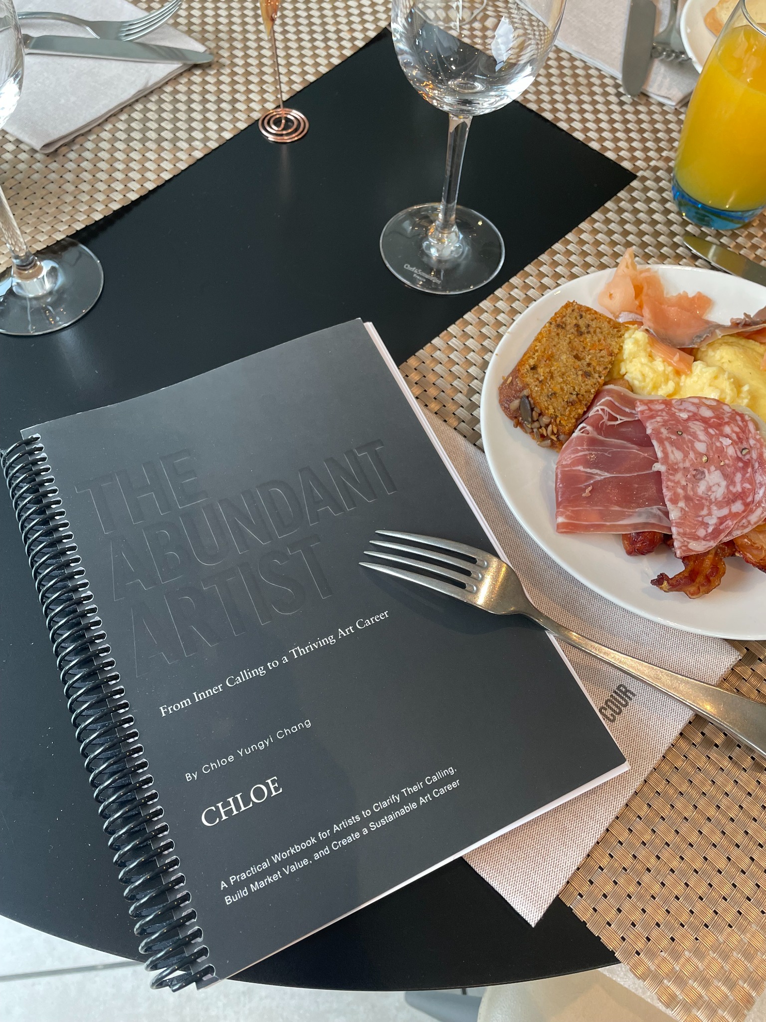 The physical, black spiral-bound workbook on a café table beside a meal